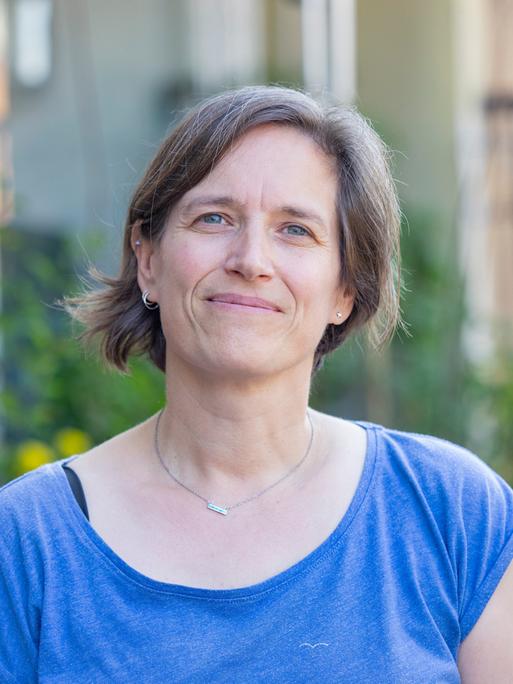 Die Traumatherapeutin Verena Fink steht vor einem verschwommenen grünen Hintergrund und blickt den Betrachter an. Sie trägt ein blaues T-Shirt und eine Halskette und hat halblange braune Haare. Die Traumatherapeutin Verena Fink steht vor einem verschwommenen grünen Hintergrund und blickt den Betrachter an. Sie trägt ein blaues T-Shirt und eine Halskette und hat halblange braune Haare.