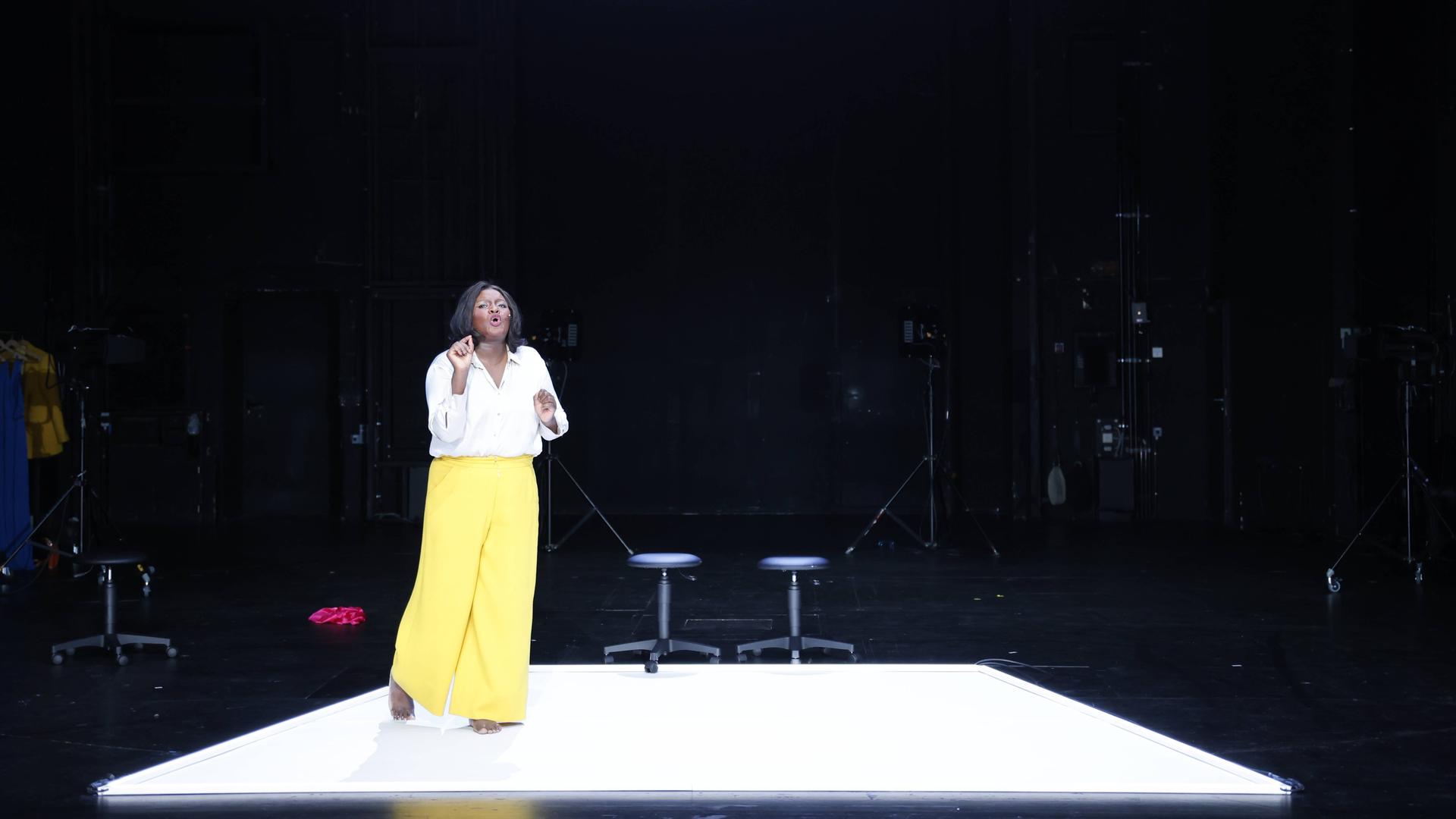 Die Schauspielerin Mercy Dorcas Otieno trägt eine gelbe Hose und ein weißes Hemd. Sie steht vor zwei Stühlen auf einer Bühne.  