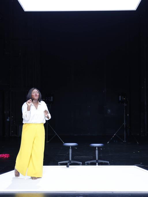 Die Schauspielerin Mercy Dorcas Otieno trägt eine gelbe Hose und ein weißes Hemd. Sie steht vor zwei Stühlen auf einer Bühne.  