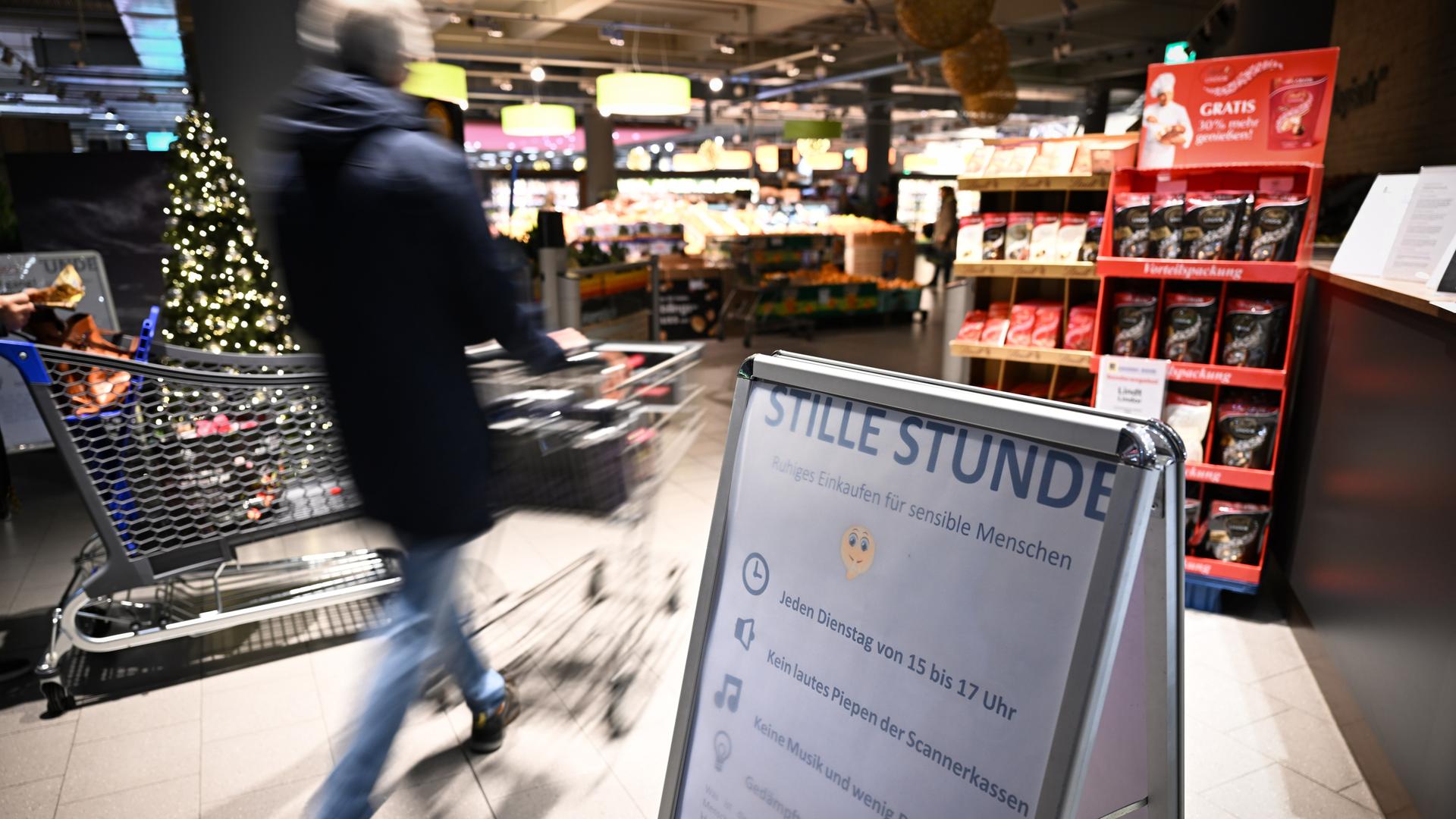 Ein Mensch schiebt einen Einkaufswagen in einen Supermarkt. Im Vordergrund steht ein Schild, das auf die "Stille Stunde" hinweist. Ein Mensch schiebt einen Einkaufswagen in einen Supermarkt. Im Vordergrund steht ein Schild, das auf die "Stille Stunde" hinweist.