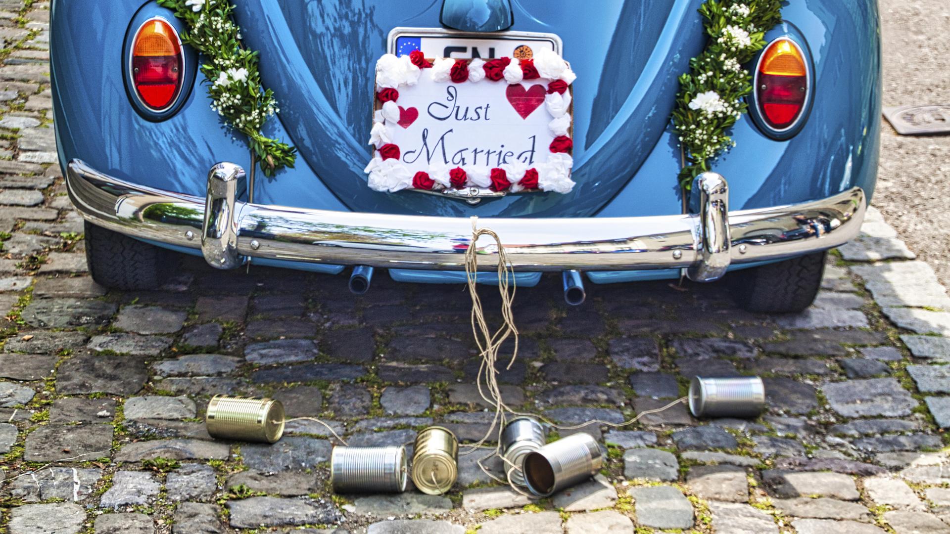 Ein Schild mit der Aufschrift „Just Married“ hängt auf der Rückseite eines blauen VW-Käfers und verdeckt das Nummernschild. An den Stoßstange sind Blechdosen an einem Seil angebracht, die auf dem Boden liegen. 