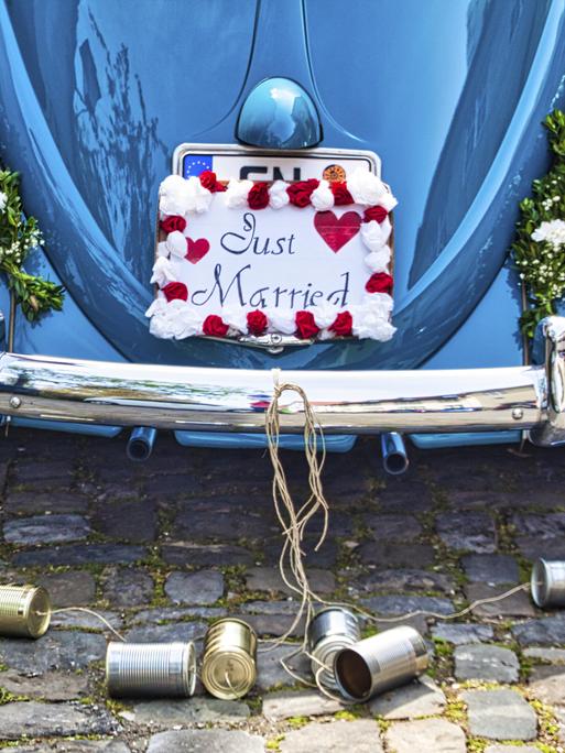 Ein Schild mit der Aufschrift „Just Married“ hängt auf der Rückseite eines blauen VW-Käfers und verdeckt das Nummernschild. An den Stoßstange sind Blechdosen an einem Seil angebracht, die auf dem Boden liegen. 