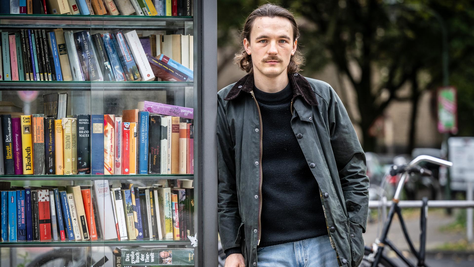 Kaleb Erdmann trägt eine schwarze Lederjacke zur Jeans und steht auf der Straße an eine alte Telefonzelle voller Bücher gelehnt. Kaleb Erdmann trägt eine schwarze Lederjacke zur Jeans und steht auf der Straße an eine alte Telefonzelle voller Bücher gelehnt.