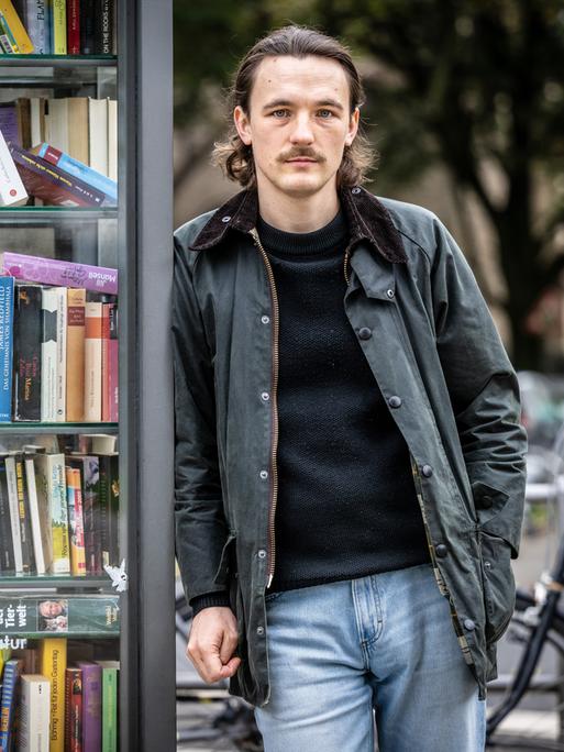 Kaleb Erdmann trägt eine schwarze Lederjacke zur Jeans und steht auf der Straße an eine alte Telefonzelle voller Bücher gelehnt. Kaleb Erdmann trägt eine schwarze Lederjacke zur Jeans und steht auf der Straße an eine alte Telefonzelle voller Bücher gelehnt.