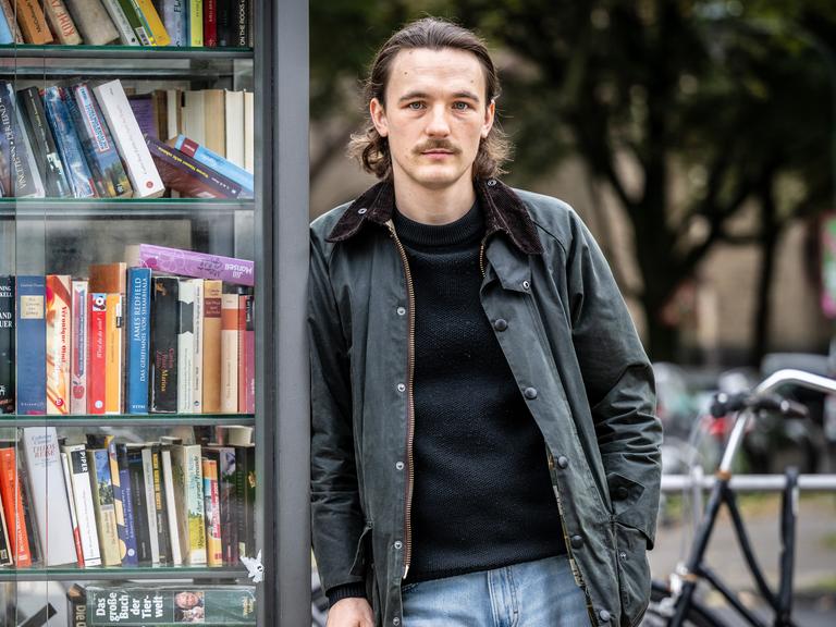 Kaleb Erdmann trägt eine schwarze Lederjacke zur Jeans und steht auf der Straße an eine alte Telefonzelle voller Bücher gelehnt. Kaleb Erdmann trägt eine schwarze Lederjacke zur Jeans und steht auf der Straße an eine alte Telefonzelle voller Bücher gelehnt.
