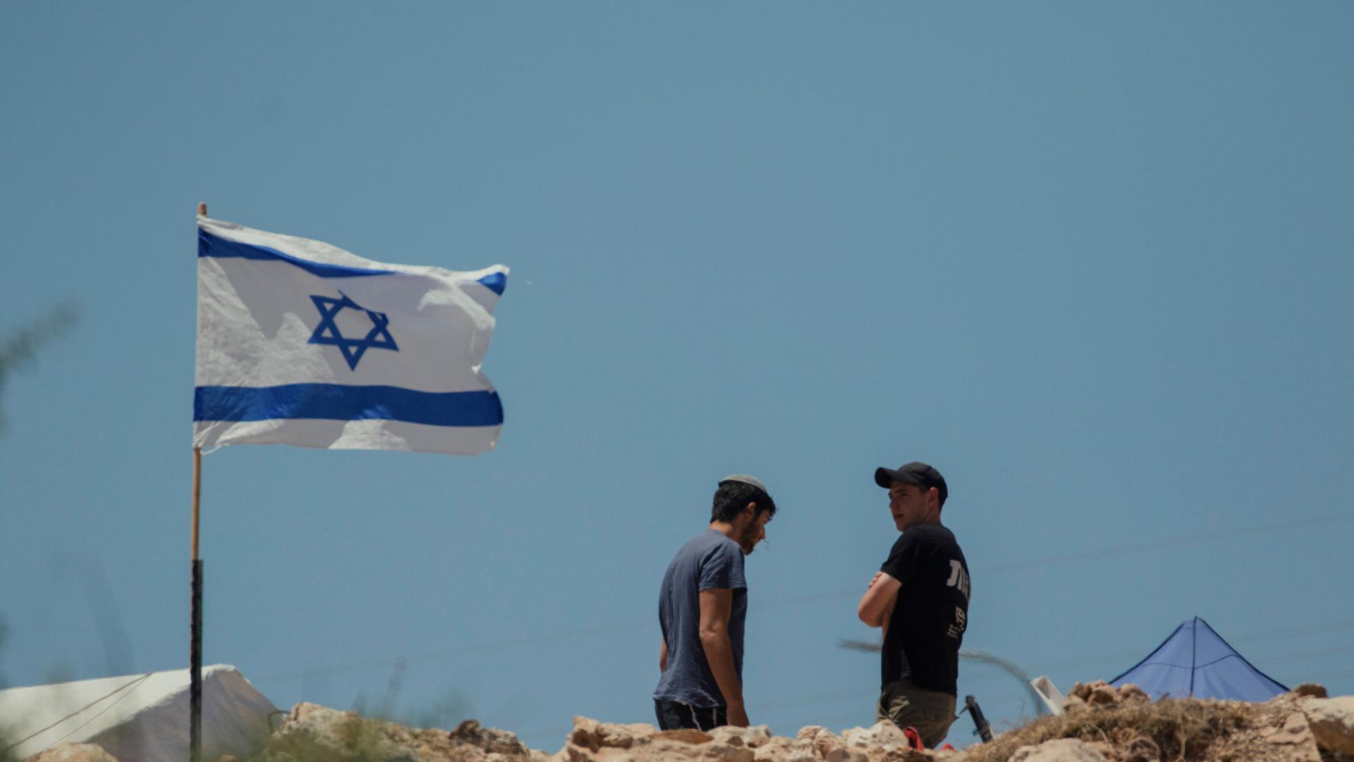 Zwei Männer stehen neben einer israelischen Flagge, die im Wind flattert.