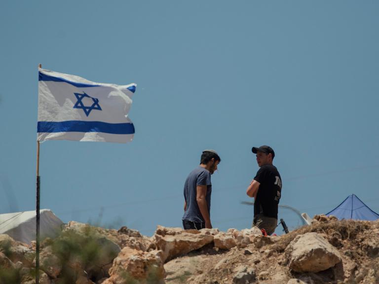 Zwei Männer stehen neben einer israelischen Flagge, die im Wind flattert.