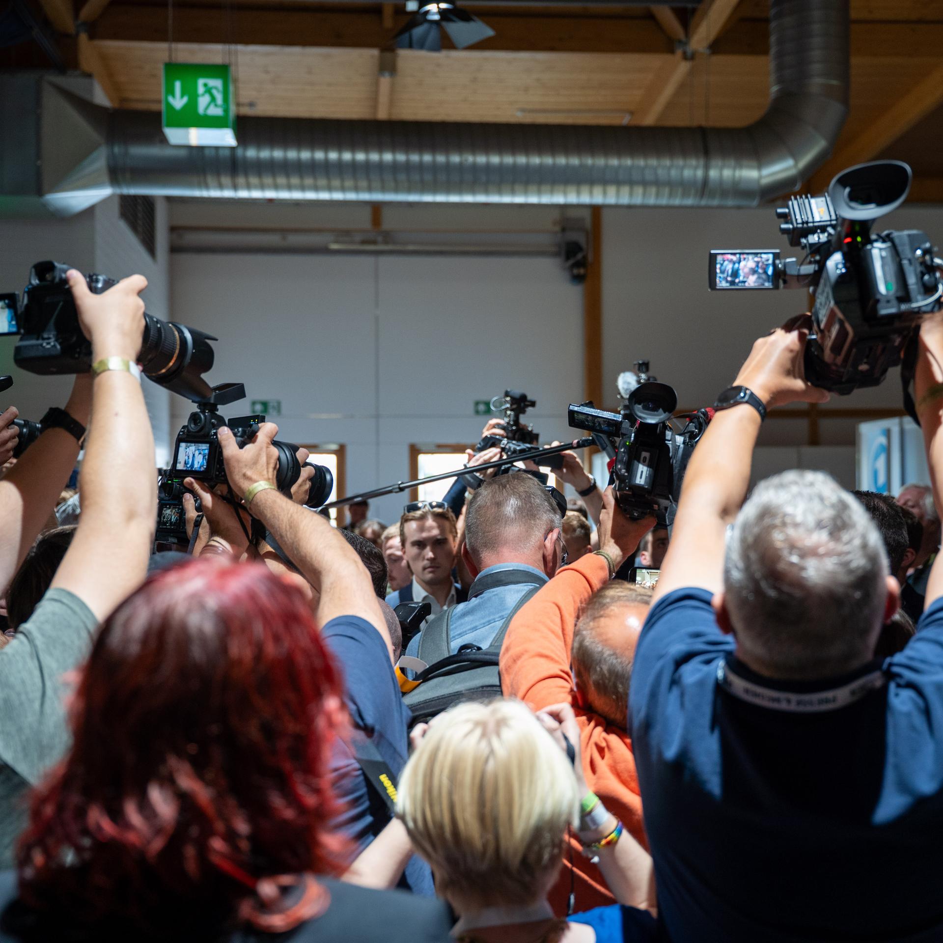 Zahlreiche Journalistinnen und Journalisten stehen mit dem Rücken zur Kamera. Sie halten eigene Kameras und Mikrofone in die Höhe.