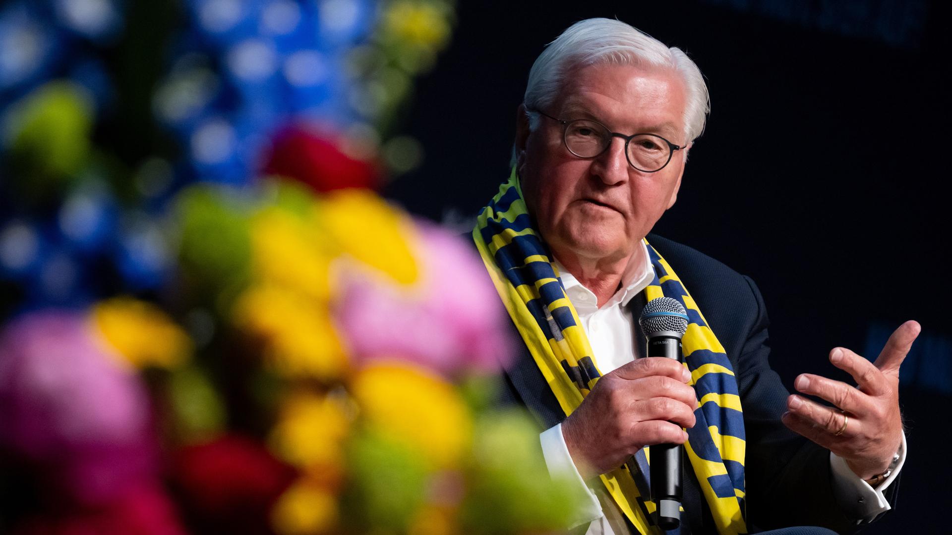 Bundespräsident Frank-Walter Steinmeier nimmt an einer Podiumsdiskussion teil. Er hat den blau-gelben Schal des Katholikentags umgelegt. Er spricht in ein Mikrofon und gestikuliert mit der Hand. 