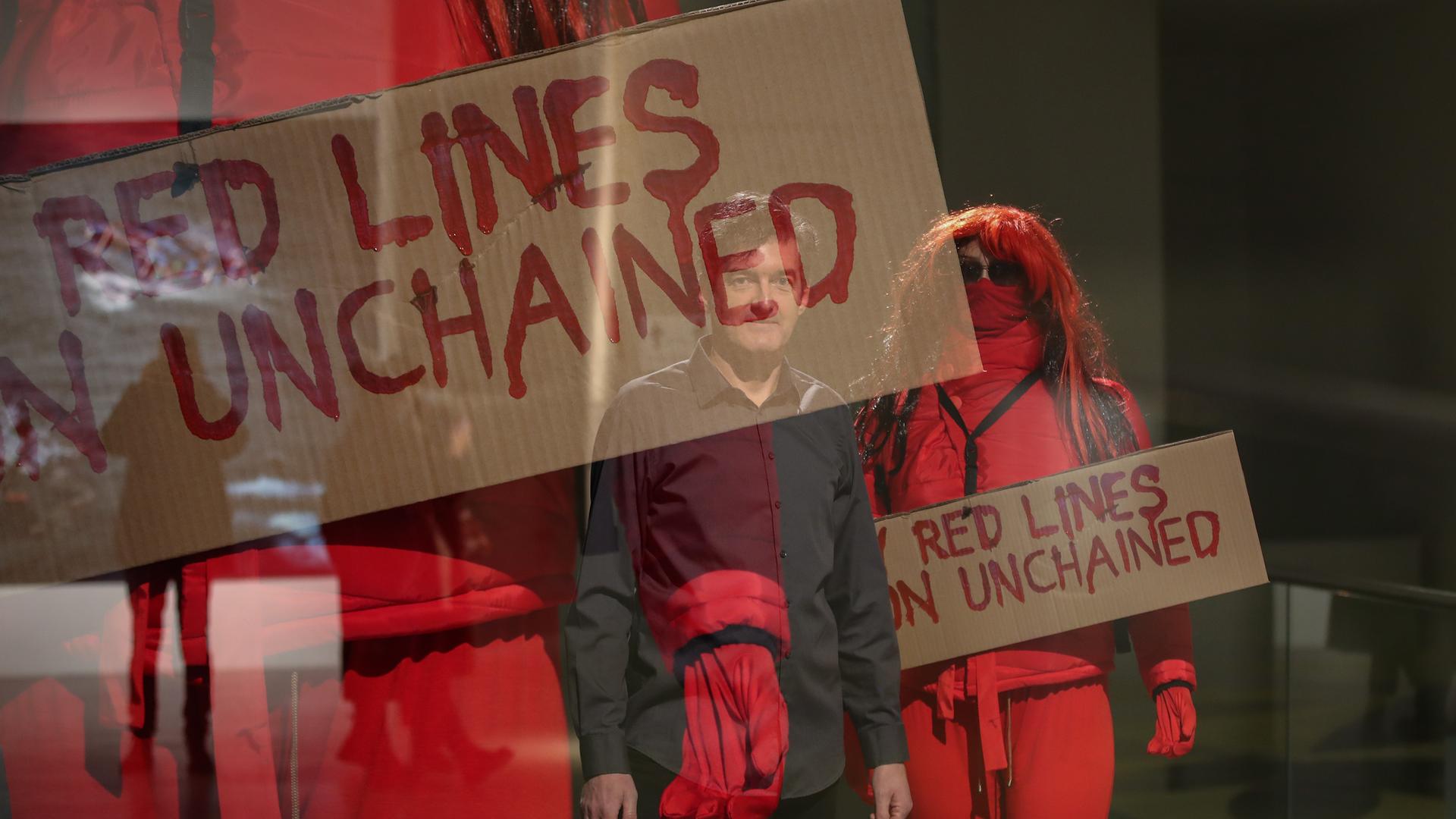 Der Videokünstler Oliver Ressler ist als Spiegelung auf einem Foto zu sehen. Dieses zeigt in rote Overalls gekleidete Aktivistinnen und Aktivisten, die Pappplakate mit der Aufschrift "Red Lines...unchained"  hochhalten.