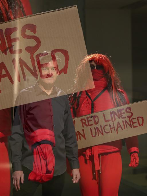 Der Videokünstler Oliver Ressler ist als Spiegelung auf einem Foto zu sehen. Dieses zeigt in rote Overalls gekleidete Aktivistinnen und Aktivisten, die Pappplakate mit der Aufschrift "Red Lines...unchained"  hochhalten.