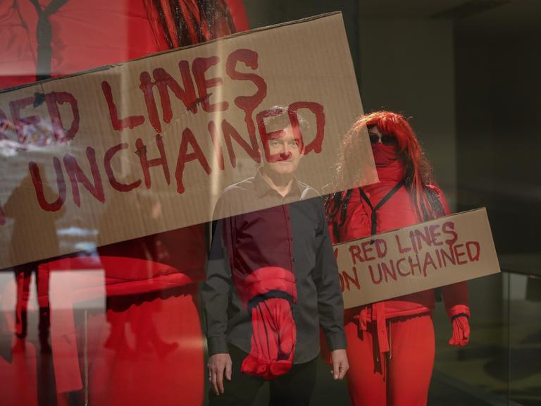 Der Videokünstler Oliver Ressler ist als Spiegelung auf einem Foto zu sehen. Dieses zeigt in rote Overalls gekleidete Aktivistinnen und Aktivisten, die Pappplakate mit der Aufschrift "Red Lines...unchained"  hochhalten.