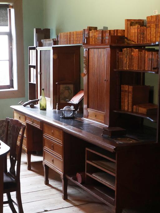 Ältere Holzmöbel stehen in einem Arbeitszimmer, am Ende des Raums fällt Licht durch ein Fenster. 