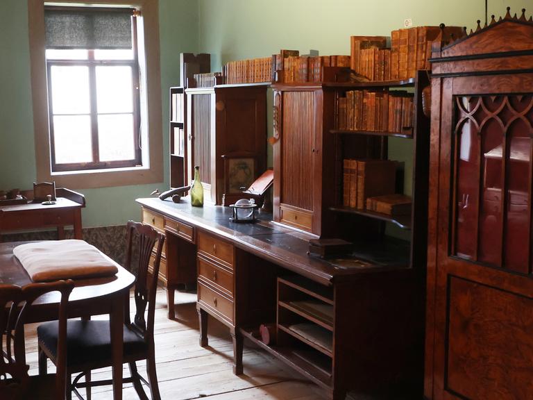 Ältere Holzmöbel stehen in einem Arbeitszimmer, am Ende des Raums fällt Licht durch ein Fenster. 