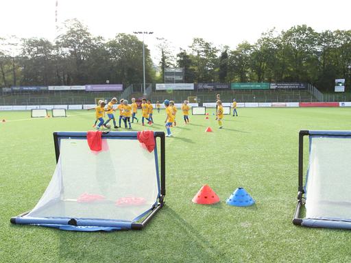 Kinder in gelben Trikots während eines Fußballtrainings auf einem Sportplatz. Im Vordergrund stehen zwei kleine Tore.