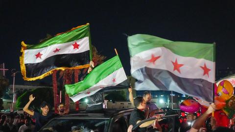 Feiernde schwenken nachts aus einem fahrenden Auto syrische Fahnen.