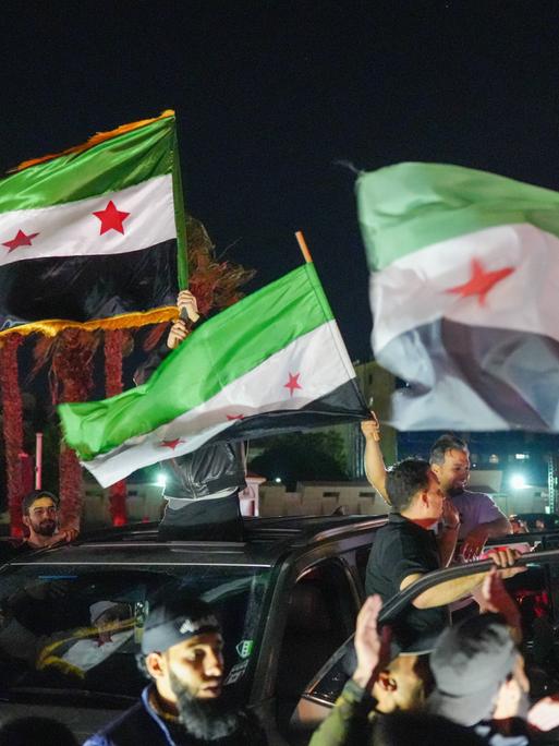 Feiernde schwenken nachts aus einem fahrenden Auto syrische Fahnen.