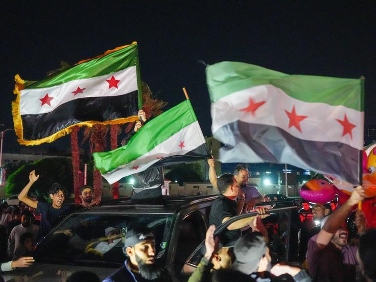 Feiernde schwenken nachts aus einem fahrenden Auto syrische Fahnen.