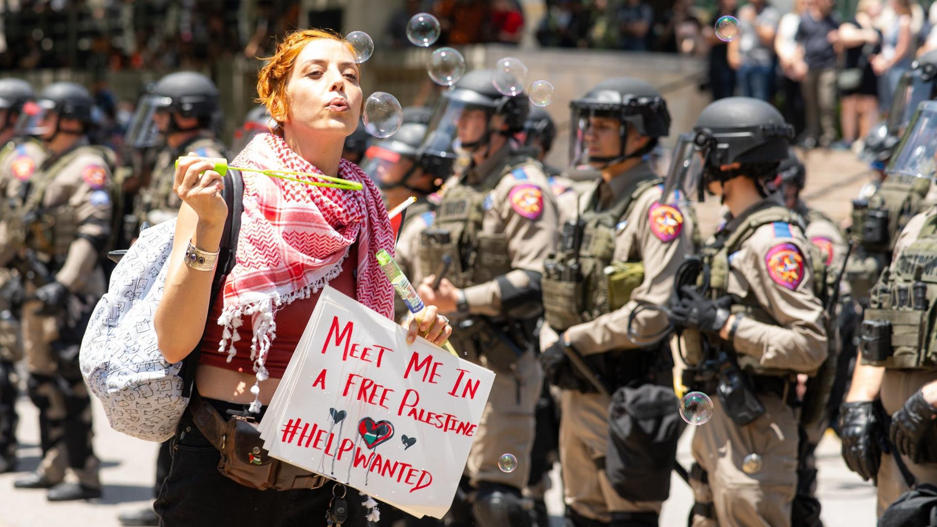Eine junge Frau bläst Seifenblasen. Sie trägt ein Protest-Plakat. Im Hintergrund sind Sicherheits-Kräfte mit Waffen. 
