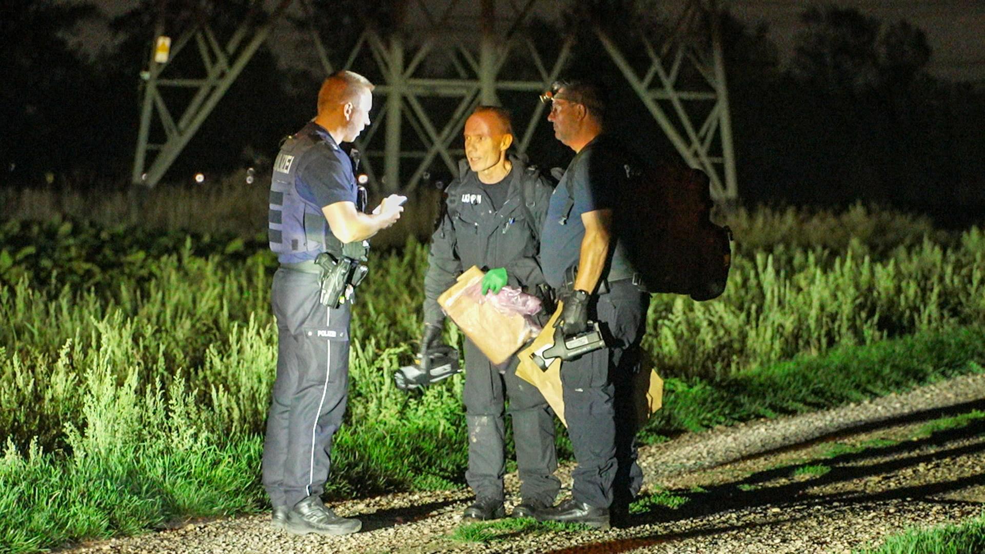 Drei Polizisten steht in der Nacht auf einem feldweg vor einem Strommast. Die Szene wird von einem Scheinwefer beleuchtet. Drei Polizisten steht in der Nacht auf einem feldweg vor einem Strommast. Die Szene wird von einem Scheinwefer beleuchtet.