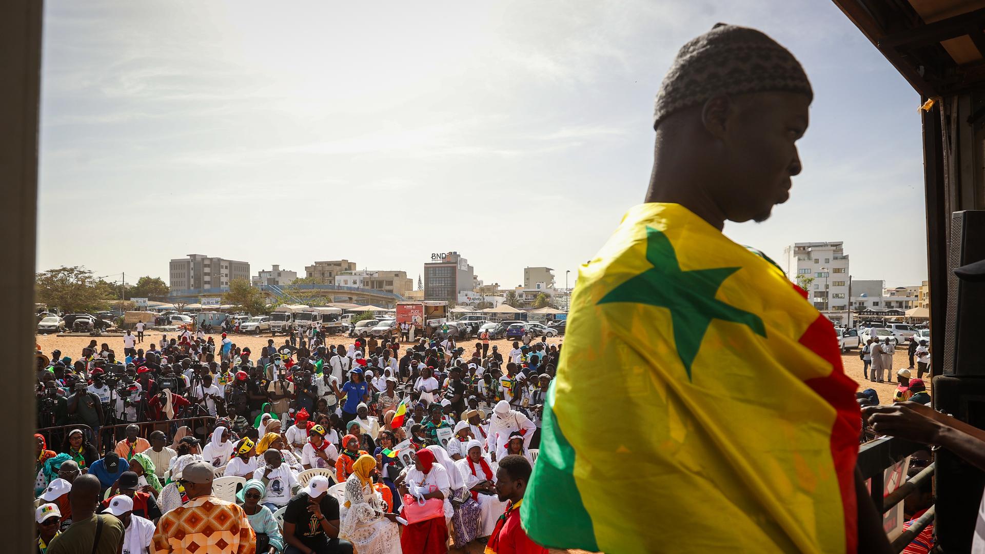  Im Bildvordergrund ist ein junger Mann eingehüllt in die senegalesische Flagge zu sehen. Im Hintergrund die von zivilgesellschaftlichen Organisationen einberufene Demonstration, auf der viele Menschen gegen die Entscheidung von Präsident Macky Sall, die Wahlen zu verschieben protestieren.

