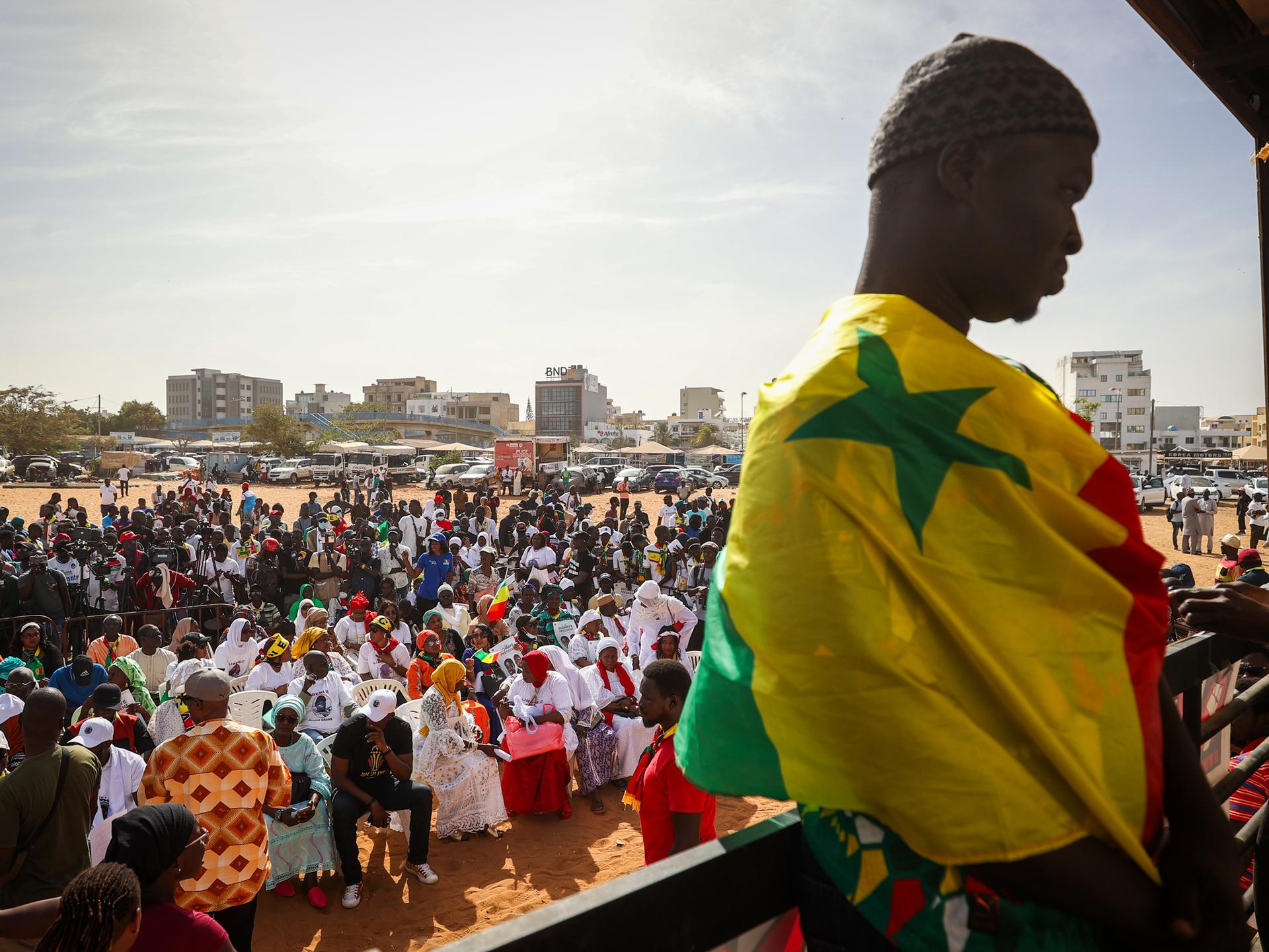  Im Bildvordergrund ist ein junger Mann eingehüllt in die senegalesische Flagge zu sehen. Im Hintergrund die von zivilgesellschaftlichen Organisationen einberufene Demonstration, auf der viele Menschen gegen die Entscheidung von Präsident Macky Sall, die Wahlen zu verschieben protestieren.

