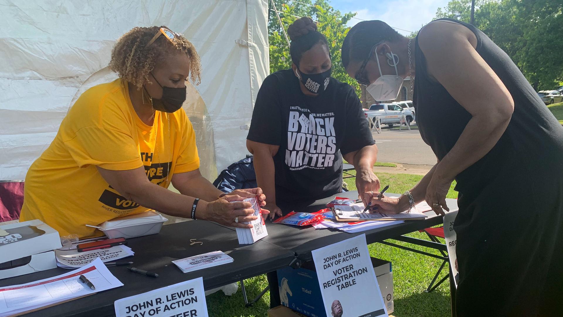Die schwarze Frau im schwarzen Kleid und mit Maske unterzeichnet ihre Registrierung an einem Stand im Freien. Hinter dem Tisch stehen zwei weitere schwarze Frauen. Sie tragen Shirts mit der Aufschrift "Black Voters Matter".