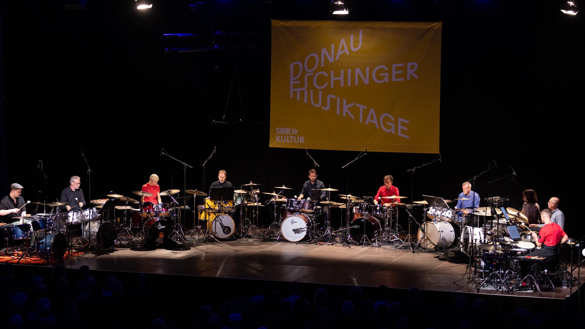 Zehn Drumsets im Halbkreis auf der Bühne, die Spieler tragen bunte einfarbige Hemden