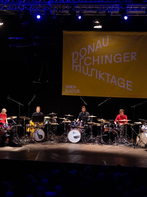 Zehn Drumsets im Halbkreis auf der Bühne, die Spieler tragen bunte einfarbige Hemden