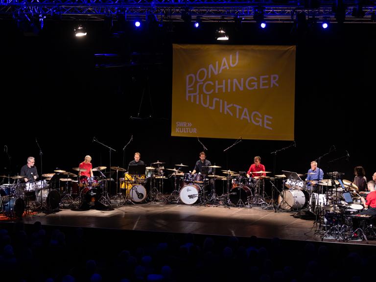 Zehn Drumsets im Halbkreis auf der Bühne, die Spieler tragen bunte einfarbige Hemden