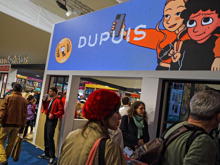 Messestände und Besucherinnen und Besucher beim Comic-Festival in Angoulême 2025. Auf einem Werbebanner sind zwei Comic-Figuren zu sehen