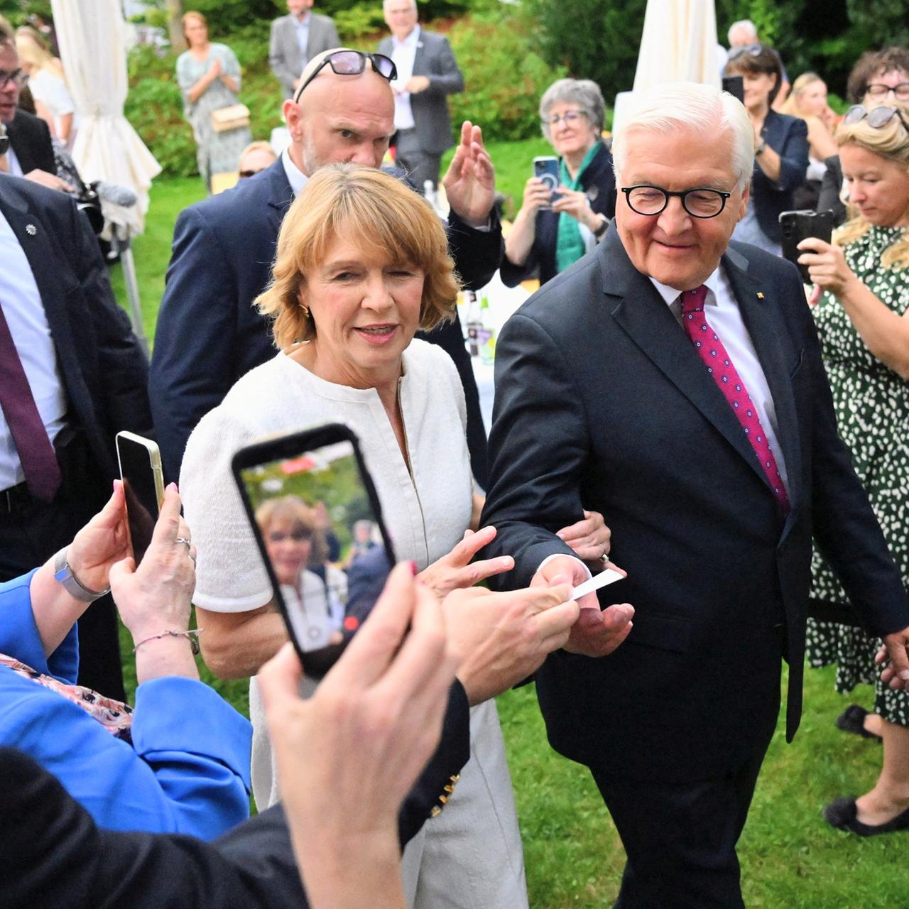Ein älteres Paar läuft über eine Wiese. Es wird umringt von Security und Menschen, die Fotos von ihnen mit ihren Handys machen.