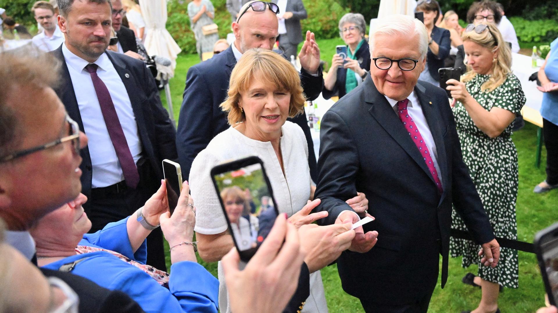 Ein älteres Paar läuft über eine Wiese. Es wird umringt von Security und Menschen, die Fotos von ihnen mit ihren Handys machen.