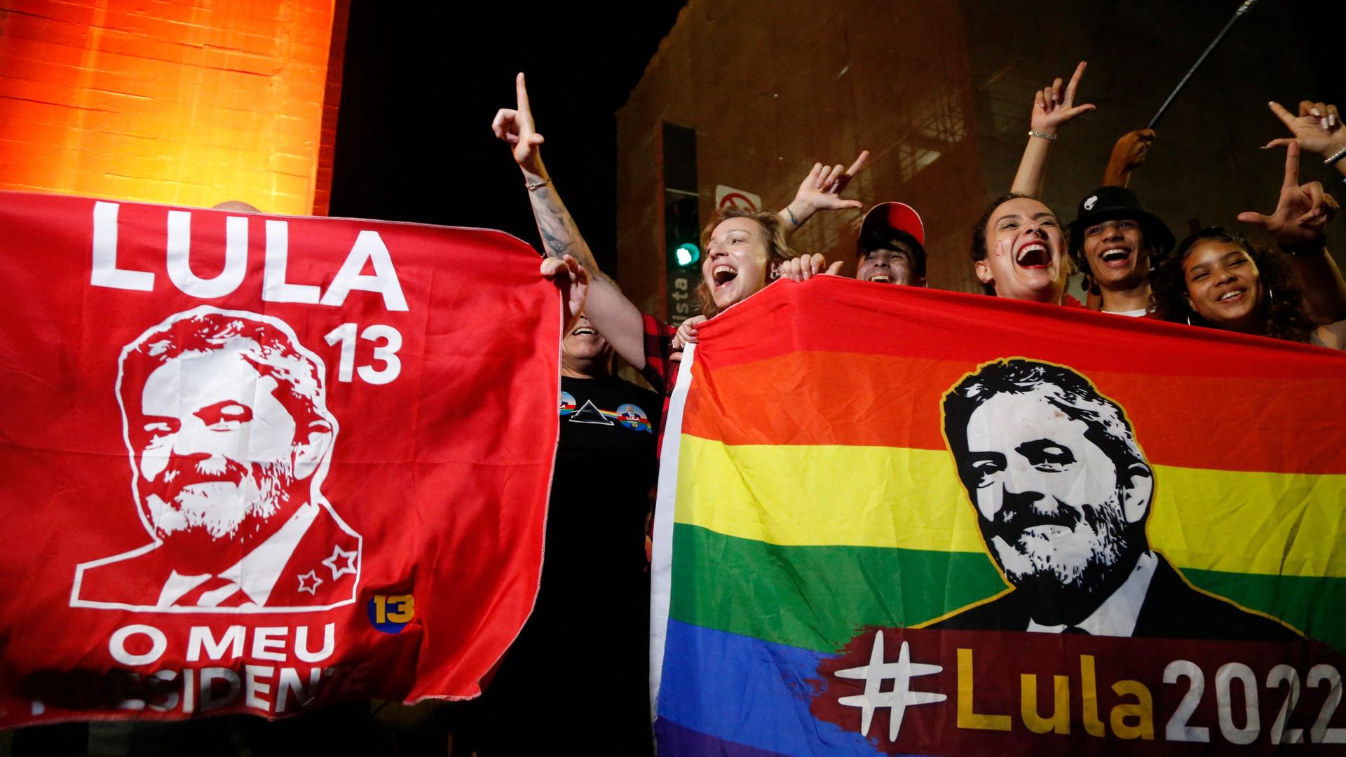 Zahlreiche Menschen feiern den Wahlsieg Lula da Silvas in Sao Paolo, Brasilien. Es ist dunkel, sie halten zwei bunte Flaggen mit dem Portrait Lulas hoch und sind fröhlich.
