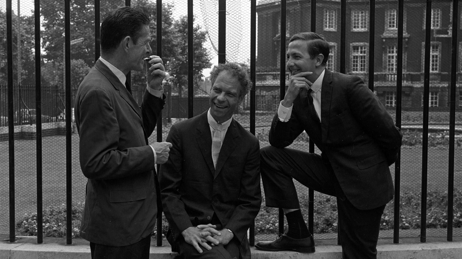 John Cage, Merce Cunningham und Robert Rauschenberg posieren, in korrekte Anzüge gekleidet, lässig vor einem Zaun beim Sadler's Wells Theater in London.
