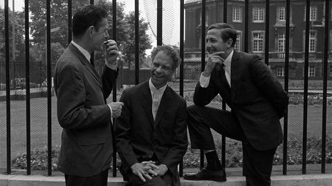 John Cage, Merce Cunningham und Robert Rauschenberg posieren, in korrekte Anzüge gekleidet, lässig vor einem Zaun beim Sadler's Wells Theater in London.