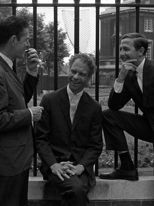 John Cage, Merce Cunningham und Robert Rauschenberg posieren, in korrekte Anzüge gekleidet, lässig vor einem Zaun beim Sadler's Wells Theater in London.