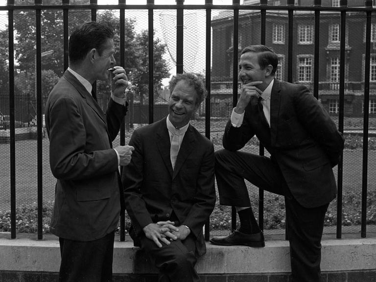 John Cage, Merce Cunningham und Robert Rauschenberg posieren, in korrekte Anzüge gekleidet, lässig vor einem Zaun beim Sadler's Wells Theater in London.