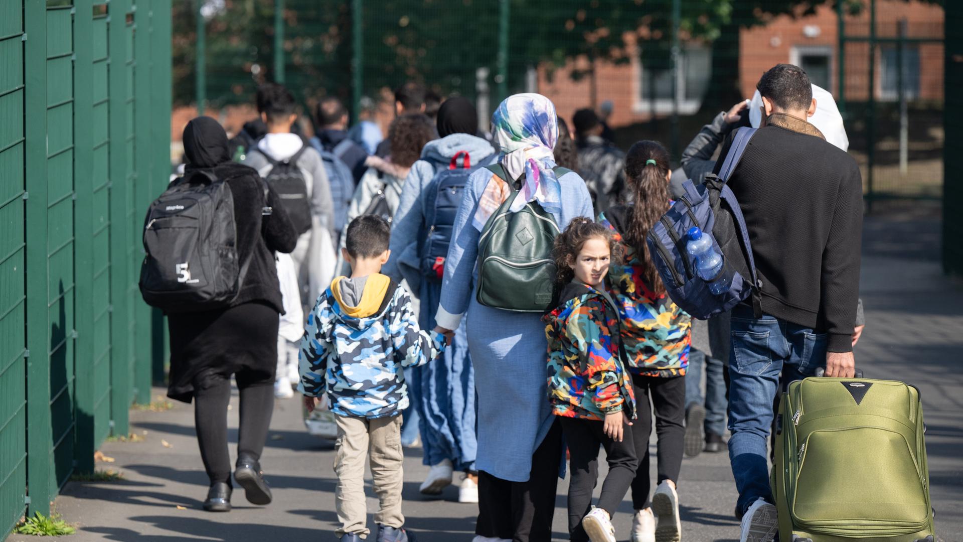 Eltern mit kleinen Kindern an den Händen gehen eine Straße entlang und ziehen einen Koffer auf Rollen