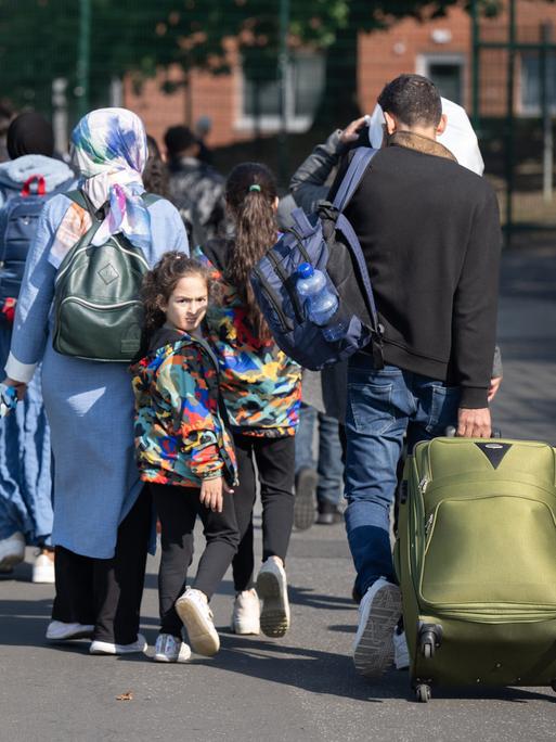 Eltern mit kleinen Kindern an den Händen gehen eine Straße entlang und ziehen einen Koffer auf Rollen