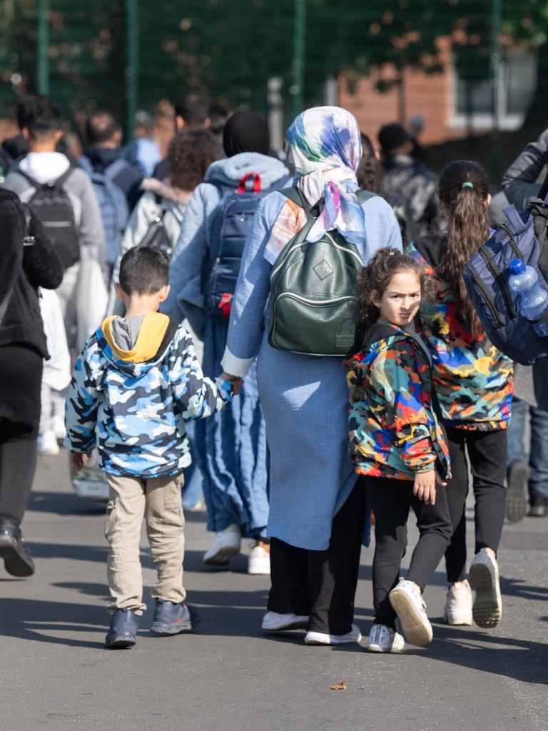Eltern mit kleinen Kindern an den Händen gehen eine Straße entlang und ziehen einen Koffer auf Rollen Eltern mit kleinen Kindern an den Händen gehen eine Straße entlang und ziehen einen Koffer auf Rollen