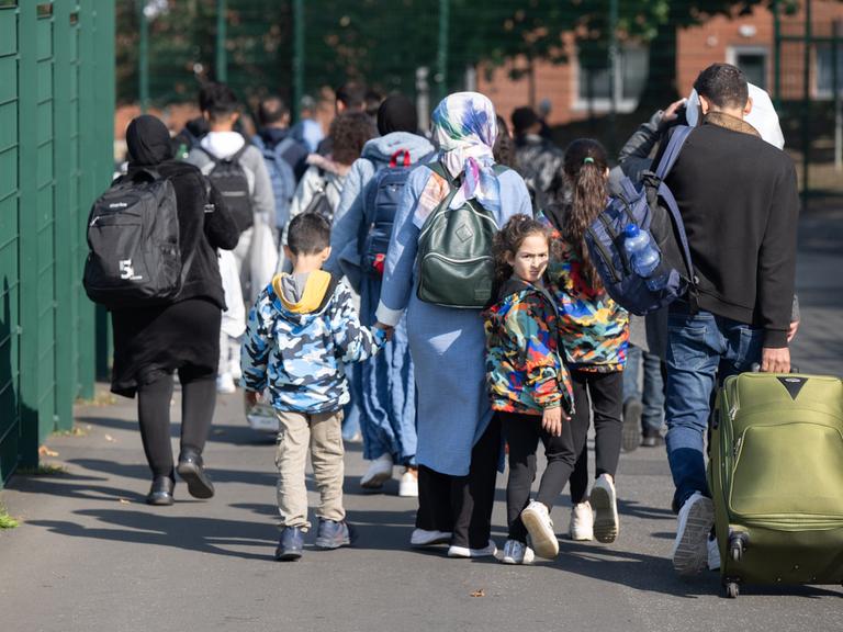 Eltern mit kleinen Kindern an den Händen gehen eine Straße entlang und ziehen einen Koffer auf Rollen