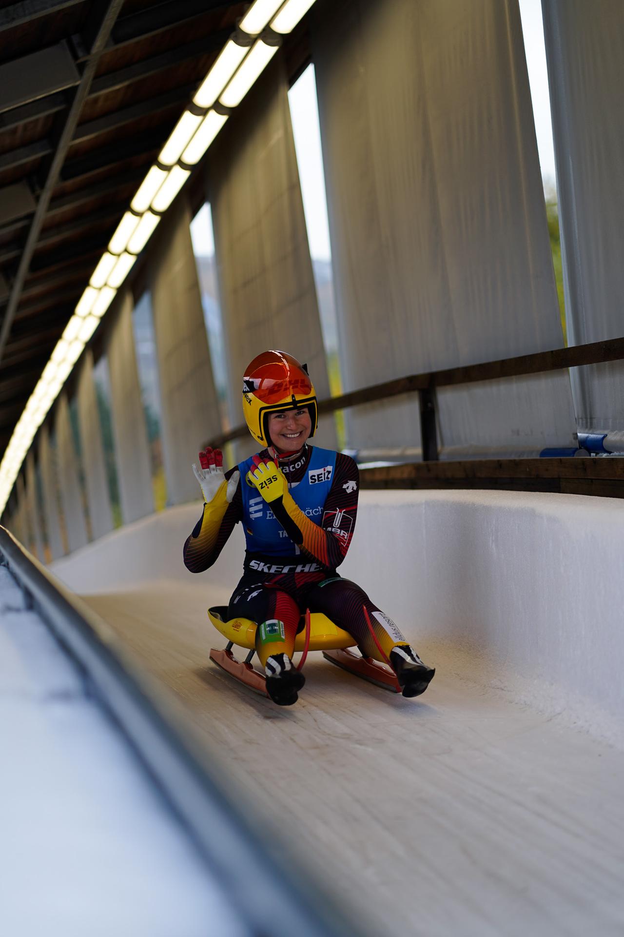Eine Rennrodlerin im bunten Sportanzug und Helm im Eiskanal. Sie lächelt in die Kamera und hält die beiden Hände so, als würde sie Querflöte spielen. Eine Rennrodlerin im bunten Sportanzug und Helm im Eiskanal. Sie lächelt in die Kamera und hält die beiden Hände so, als würde sie Querflöte spielen.