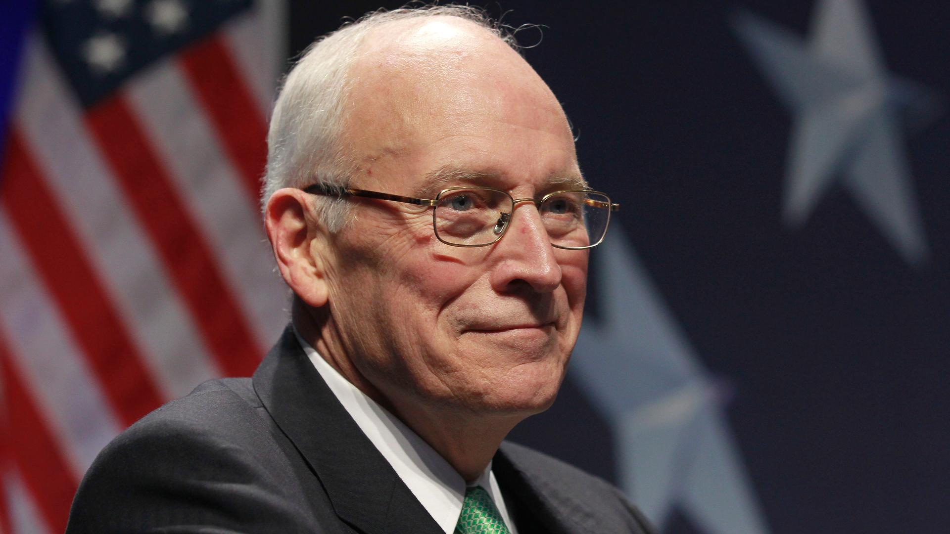 Dick Cheney trägt Brille und sitzt vor einer amerikanischen Flagge Dick Cheney trägt Brille und sitzt vor einer amerikanischen Flagge