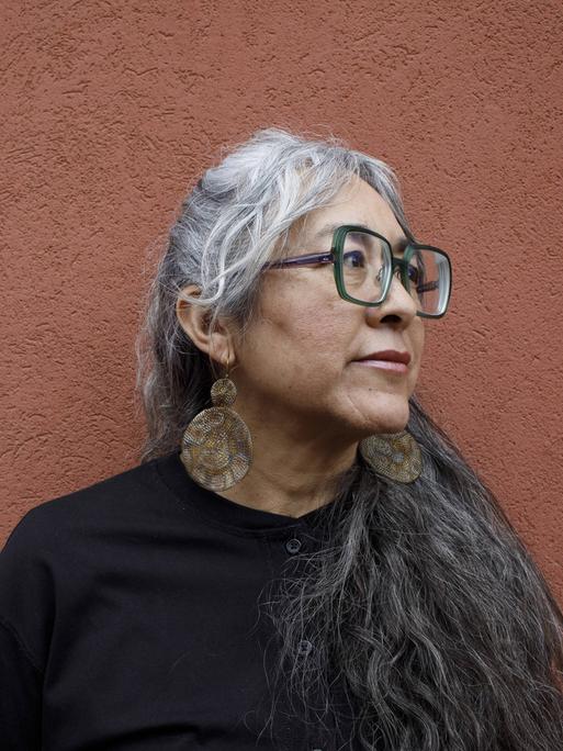 Cristina Rivera Garza steht vor einer ockerfarbenen Wand und schaut zur Seite.