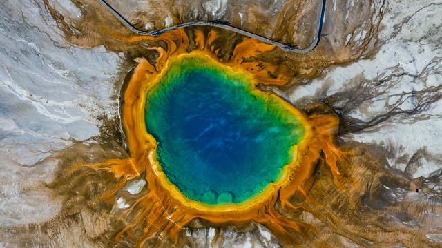 Aus der Luft aufgenommen: Ein knallblauer klarer See in felsiger Landschaft, die ihn einfasst - am Rand ist das Wasser eingefärbt mit verschiedenen Streifen in Regenbogenfarben.