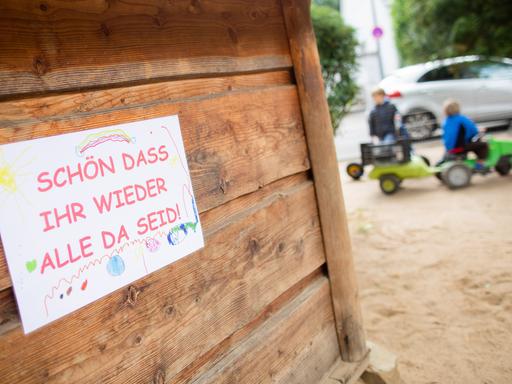 Kinder spielen auf dem Spielplatz einer Kindertagesstätte, ein Schild "Schön dass ihr wieder alle da seid" hängt an einer Holzwand. (Coronavirus)