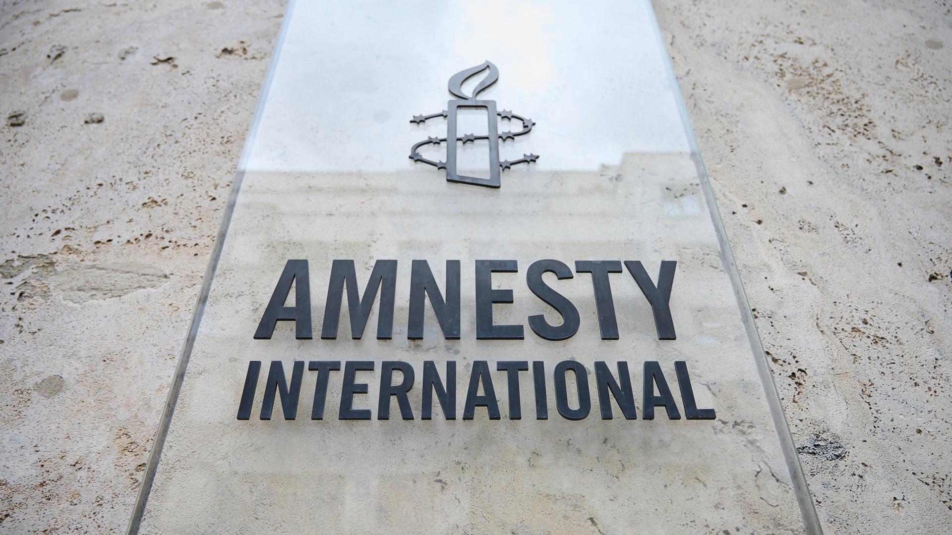 Das Logo von Amnesty International auf einer transparenten Schild an einer Fassade in Berlin. Es zeigt eine brennende Kerze, die mit Stacheldraht umwickelt ist.