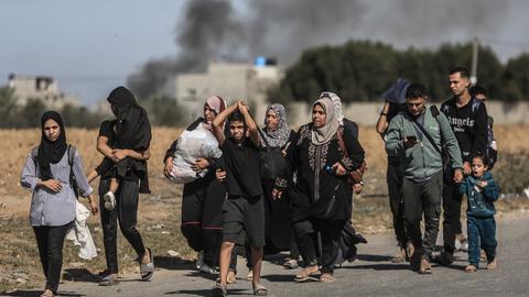 Palästinensische Kinder, Frauen und Männer auf der Flucht im Gazastreifen. Manche der Kinder werden von anderen getragen. Einer der Männer blickt auf sein Handy. Ein kleines Mädchen gähnt. Die Gruppe hat wenig Gepäck. Im Hintergrund steigt Rauch auf.
