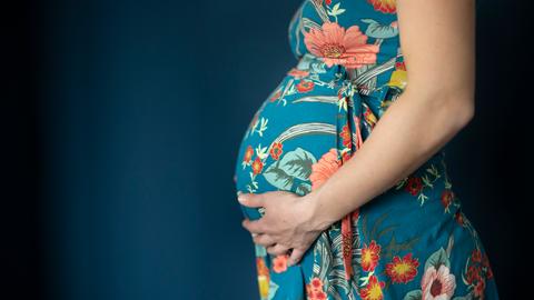 Eine junge Frau trägt ein blaues Kleid mit Blumen und hält mit einer Hand ihren runden Schwangerschaftsbauch.